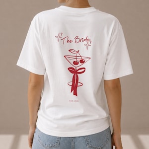 Op de afbeelding: Een wit T-shirt met korte mouwen en een rode afbeelding op de achterkant. Het ontwerp bevat de tekst "The Bride" boven een martiniglas met twee kersen en een strik, met "EST. 2026" onder het glas en vierpuntige sterren als accenten.