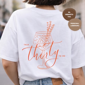 Può includere: T-shirt bianco con un disegno arancione di un bicchiere da cocktail, una fetta di limone, una cannuccia e la scritta "Talk Thirty to Me". Il disegno comprende rametti di erbe. La maglietta è abbinata a jeans blu.