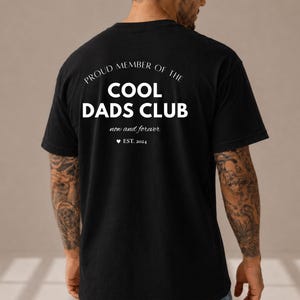 Könnte beinhalten: Schwarzes T-Shirt mit weißem Aufdruck "PROUD MEMBER OF THE COOL DADS CLUB", darunter "now and forever" und "EST. 2024". Das Shirt hat einen Rundhalsausschnitt.