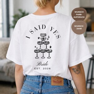 May include: White t-shirt featuring a black graphic on the back. The design includes the text "I SAID YES" above a champagne glass tower, the word "Bride", and the year "EST. 2026".