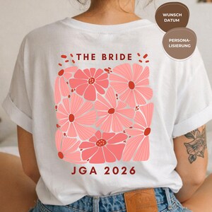 Könnte beinhalten: Weißes T-Shirt mit einem rosa Blumendesign. Der Text "THE BRIDE" befindet sich über den Blumen und "JGA 2026" darunter. Das T-Shirt hat hochgekrempelte Ärmel.