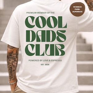 May include: Cream-colored t-shirt with the text "COOL DADS CLUB" in dark green, with the words "PREMIUM MEMBER OF THE" above and "POWERED BY LOVE & ESPRESSO EST. 2026" below. A brown circle with German text is visible.