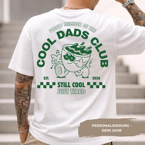 Può includere: T-shirt bianco con grafica verde sul retro. Il design include il testo "COOL DADS CLUB" attorno a un personaggio di tazza di caffè dei cartoni animati, con "EST. 2026" e "STILL COOL JUST TIRED" sotto.