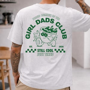 May include: Light gray t-shirt with a green graphic on the back. The graphic reads "GIRL DADS CLUB" with a cartoon character and the text "EST. 2026 STILL COOL JUST TIRED".