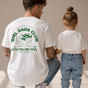 May include: White t-shirt with green text and graphic. The text reads "GIRL DADS CLUB" and "STILL COOL JUST TIRED". A person wearing the shirt is kneeling next to a child. The graphic is a cartoon of a cup with sunglasses.