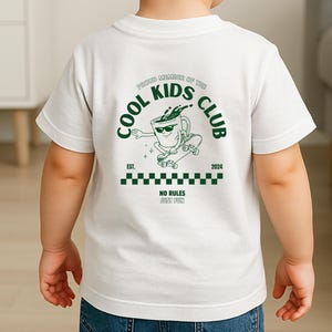 May include: White t-shirt with a green graphic on the back. The graphic features a coffee cup character skateboarding, with the text "COOL KIDS CLUB" and "NO RULES JUST FUN". The shirt is worn by a child.