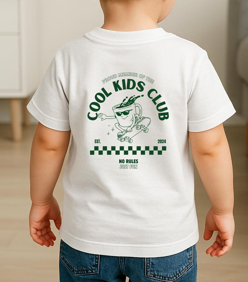 May include: White t-shirt with a green graphic on the back. The graphic features a coffee cup character skateboarding, with the text "COOL KIDS CLUB" and "NO RULES JUST FUN". The shirt is worn by a child.