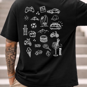 May include: A black t-shirt featuring a white graphic print on the back. The design consists of various line art icons including a game controller, car, surfboard, van, bicycle, burger, ice cream, dumbbell, and the text DAD and 1998.