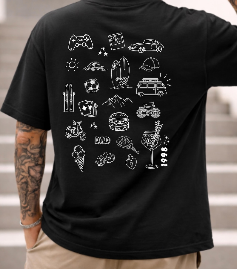 May include: A black t-shirt featuring a white graphic print on the back. The design consists of various line art icons including a game controller, car, surfboard, van, bicycle, burger, ice cream, dumbbell, and the text DAD and 1998.