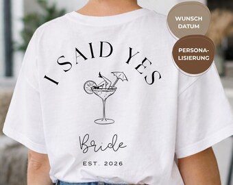 May include: White t-shirt featuring the text "I Said Yes" in black, curved across the top. A cocktail glass graphic with a person relaxing inside is below. The word "Bride" is written in cursive, with "EST. 2026" underneath.