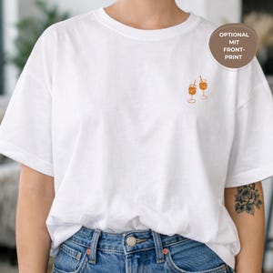 May include: White t-shirt with a small embroidered design of two orange cocktails in stemmed glasses. The shirt is a relaxed fit and is paired with blue denim jeans. A circular label reads "OPTIONAL MIT FRONT-PRINT".