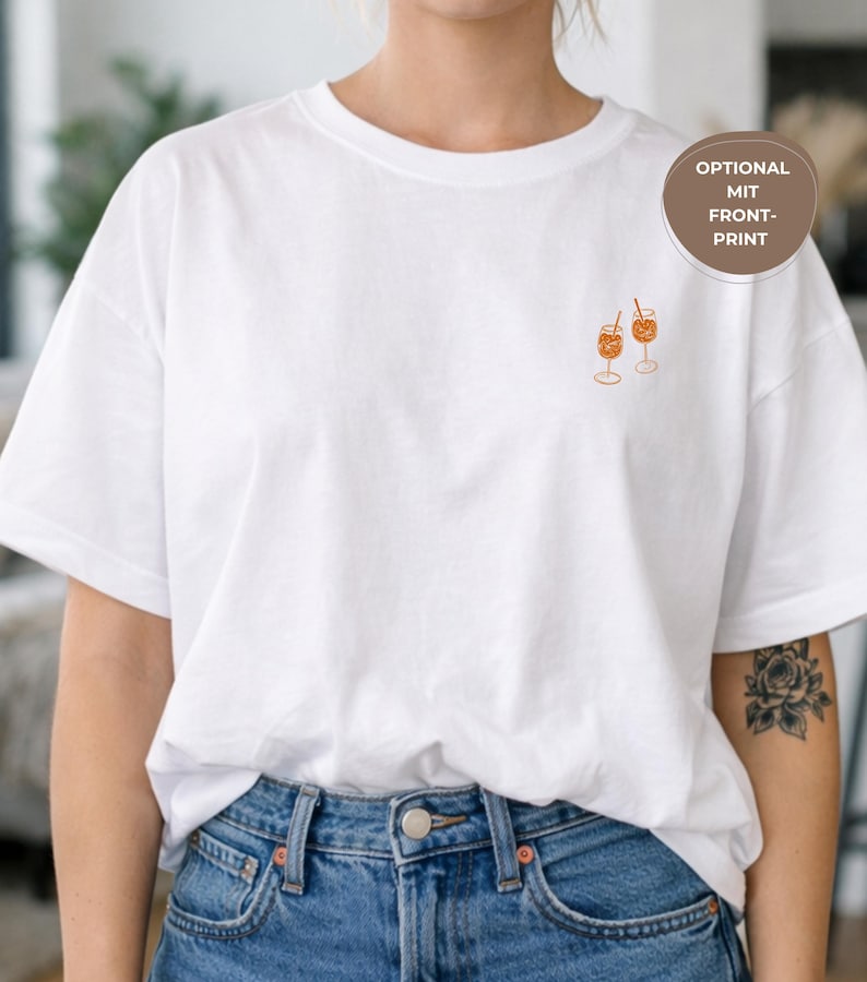 May include: White t-shirt with a small embroidered design of two orange cocktails in stemmed glasses. The shirt is a relaxed fit and is paired with blue denim jeans. A circular label reads "OPTIONAL MIT FRONT-PRINT".
