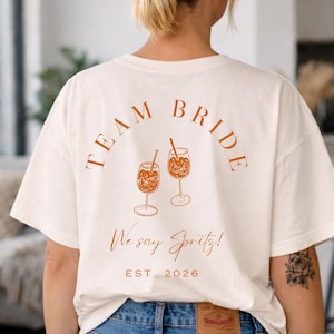 May include: Cream-colored t-shirt with the words "TEAM BRIDE" arched across the top in a burnt orange color. Below are two illustrations of cocktails, and the text "We say Spritz!" and "EST. 2026" in the same color.