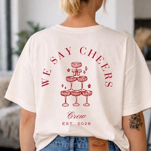 May include: A cream-coloured t-shirt featuring a red graphic of champagne glasses arranged in a pyramid, with the words "WE SAY CHEERS" above. The words "Crew" and "EST. 2026" are below the graphic.