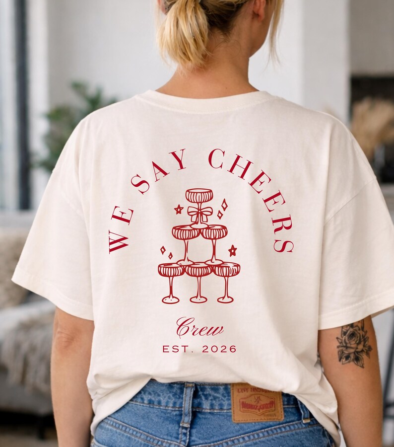 May include: A cream-coloured t-shirt featuring a red graphic of champagne glasses arranged in a pyramid, with the words "WE SAY CHEERS" above. The words "Crew" and "EST. 2026" are below the graphic.