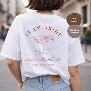 Op de afbeelding: Wit T-shirt met rode tekst en een cocktailafbeelding. De woorden "TEAM BRIDE" staan boven drie martiniglazen. Daaronder staat de tekst "Laura's Bachelorette MALLORCA 2026".