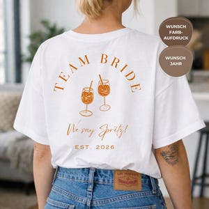 May include: White t-shirt with the words "TEAM BRIDE" in a burnt orange color, along with two spritz drink illustrations. The text "We say Spritz!" and "EST. 2026" are also printed on the shirt.