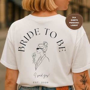 Op de afbeelding: Wit T-shirt met de woorden "BRIDE TO BE" in een boog bovenaan, en een lijntekening van een vrouw met een zonnebril en een drankje. Daaronder staat de tekst "I said yes!" en "EST. 2026".