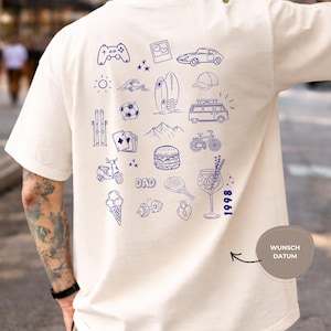 May include: An off-white t-shirt featuring a back print of various blue line-art icons including a game controller, car, surfboard, van, bicycle, burger, ice cream, and the word DAD. The number 1998 is printed vertically near the bottom right.