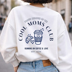 Può includere: Felpa bianca con grafica blu navy sul retro. La grafica include il testo "COOL MOMS CLUB" e "RUNNING ON COFFEE & LOVE EST. 2026" con un'illustrazione di tazza di caffè e croissant.