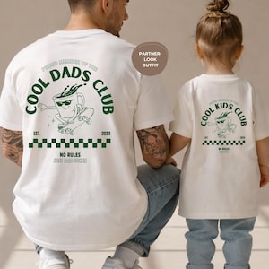 May include: White t-shirts with matching graphics. The adult shirt reads "Cool Dads Club" with a skateboarding coffee cup design. The child's shirt says "Cool Kids Club" with a similar design. Both shirts have the text "No Rules Just Dad Jokes".