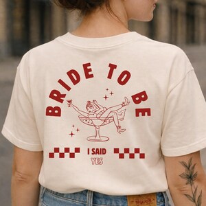 Puede incluir: Camiseta color crema con las palabras "BRIDE TO BE" en rojo arqueadas. Debajo, una ilustraci&oacute;n roja de una mujer en una copa de c&oacute;ctel, con el texto "I SAID YES". La camiseta es de estilo casual.