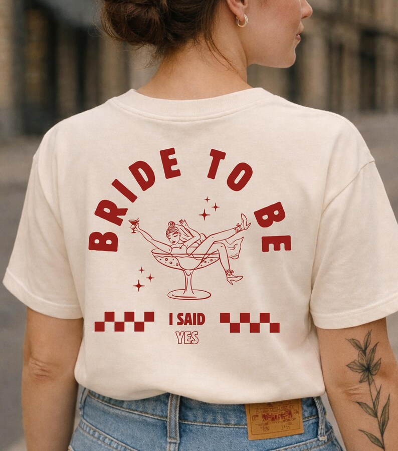 Puede incluir: Camiseta color crema con las palabras "BRIDE TO BE" en rojo arqueadas. Debajo, una ilustraci&oacute;n roja de una mujer en una copa de c&oacute;ctel, con el texto "I SAID YES". La camiseta es de estilo casual.
