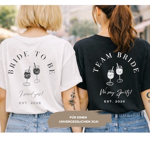 May include: Two t-shirts, one white and one black, with matching designs. The white shirt says "Bride to Be" with "I said yes!" and "EST. 2026". The black shirt says "Team Bride" with "We say Spritz!" and "EST. 2026". Both shirts feature cocktail illustrations.