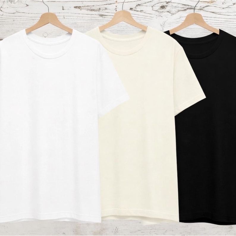 May include: Three short-sleeved t-shirts hanging on wooden hangers. The first shirt is solid white, the second shirt is a light cream or off-white colour, and the third shirt is solid black. Each shirt features a classic crew neckline and plain design.