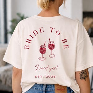 May include: Cream-colored t-shirt with the text "BRIDE TO BE" arched across the top, and two wine glasses with fruit and straws. Below the glasses, the text reads "I said yes!" and "EST. 2026".