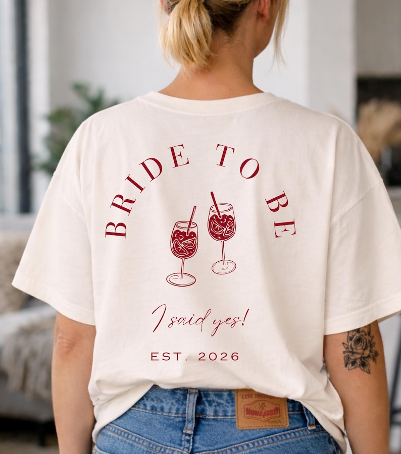 May include: Cream-colored t-shirt with the text "BRIDE TO BE" arched across the top, and two wine glasses with fruit and straws. Below the glasses, the text reads "I said yes!" and "EST. 2026".