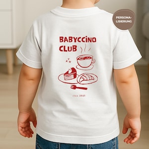 May include: White t-shirt with a red graphic on the back featuring the text "BABYCCINO CLUB" above a coffee cup, cake, and croissant. The phrase "since 2025" is printed below the illustration. The shirt is worn by a child.