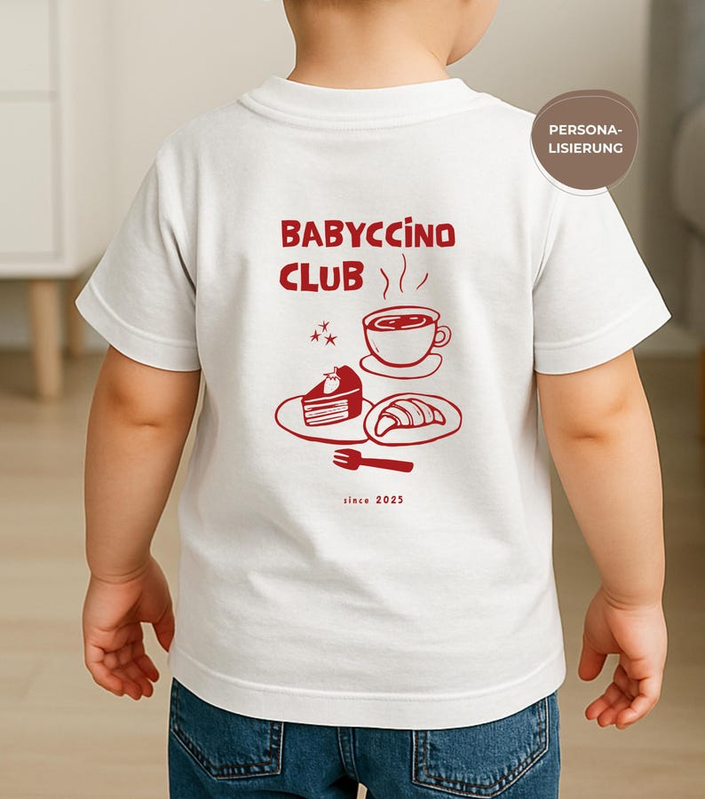 May include: White t-shirt with a red graphic on the back featuring the text "BABYCCINO CLUB" above a coffee cup, cake, and croissant. The phrase "since 2025" is printed below the illustration. The shirt is worn by a child.