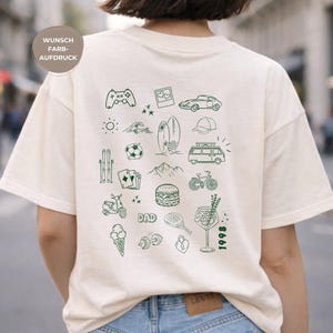 Op de afbeelding: Crèmekleurig T-shirt met een groene doodle-design. Het ontwerp bevat een gamecontroller, auto, surfboard, berg, ijsje en het woord "DAD". Het jaar "1998" is ook te zien. Het shirt is gemaakt van een zacht materiaal.