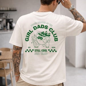 May include: White t-shirt with a green graphic on the back that reads "GIRL DADS CLUB" in a circular design. The graphic includes a cartoon image of a bowl with a face, and the text "EST. 2026 STILL COOL JUST TIRED".