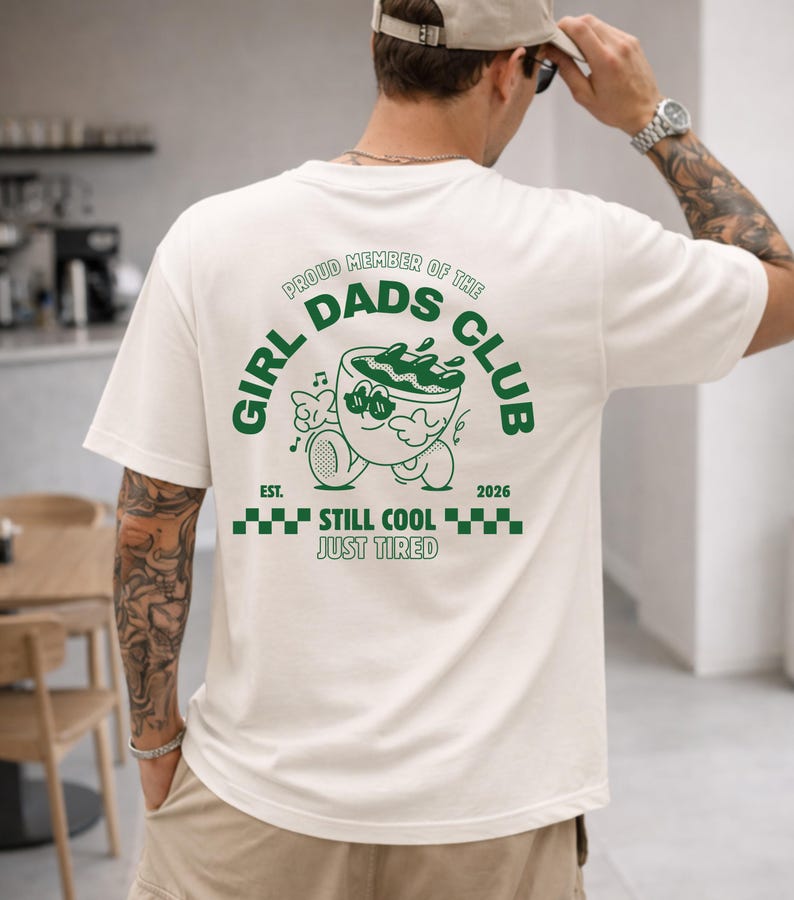 May include: White t-shirt with a green graphic on the back that reads "GIRL DADS CLUB" in a circular design. The graphic includes a cartoon image of a bowl with a face, and the text "EST. 2026 STILL COOL JUST TIRED".