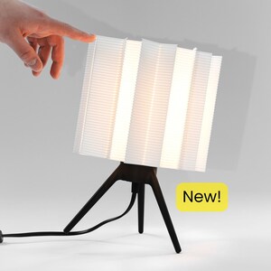 May include: A modern white table lamp with a unique, pleated shade design. The lamp has a dark brown tripod base and a cord. The image includes the text "New!" on a yellow background.
