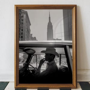 May include: Black and white framed photograph of a person in a cowboy hat smoking a cigarette while sitting in a car. The Empire State Building is visible in the background, framed by the car window.