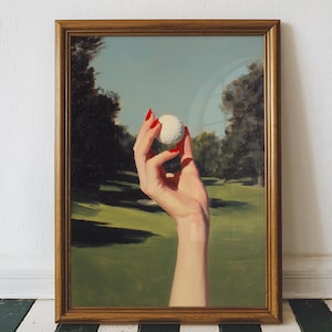 May include: A framed art print featuring a golf ball held by a hand with red nail polish. The background depicts a golf course with green grass and trees under a blue sky. The frame is a warm, golden brown.
