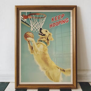 May include: A framed poster with a golden retriever jumping to dunk a basketball. The poster has a blue background with a basketball hoop and the text 'KEEP HOOPING' in red letters.