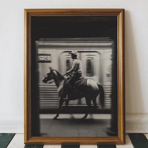 May include: Framed black and white photograph of a cowboy riding a horse in front of a speeding subway train. The image is in a gold-colored wooden frame, set against a white wall and a black and white striped floor.