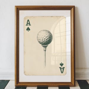 May include: A framed print of a playing card with a golf ball on a tee. The card is the ace of spades and is printed in green and white on a cream background. The card is framed in a brown wood frame.