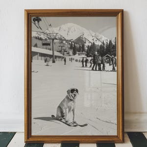 May include: Black and white framed print of a dog wearing ski goggles and skis, sitting on a snowy ski slope. The background shows a ski lift, skiers, and a mountain range. The frame is a warm brown color.