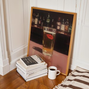 Puede incluir: Una impresi&oacute;n enmarcada de una escena de bar con un vaso de cerveza con una huella de labios rojos. Una pila de libros, una taza blanca con caf&eacute; y una alfombra a rayas est&aacute;n en el suelo. La impresi&oacute;n tiene un marco de madera.
