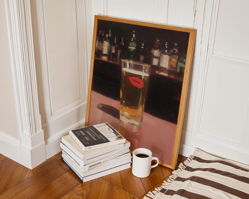 Puede incluir: Una impresi&oacute;n enmarcada de una escena de bar con un vaso de cerveza con una huella de labios rojos. Una pila de libros, una taza blanca con caf&eacute; y una alfombra a rayas est&aacute;n en el suelo. La impresi&oacute;n tiene un marco de madera.