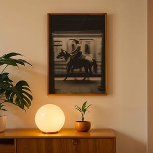 May include: A framed black and white photograph of a person riding a horse near a train. The photograph is mounted on a light-colored wall above a wooden cabinet. A glowing white orb lamp and potted plants are on the cabinet.