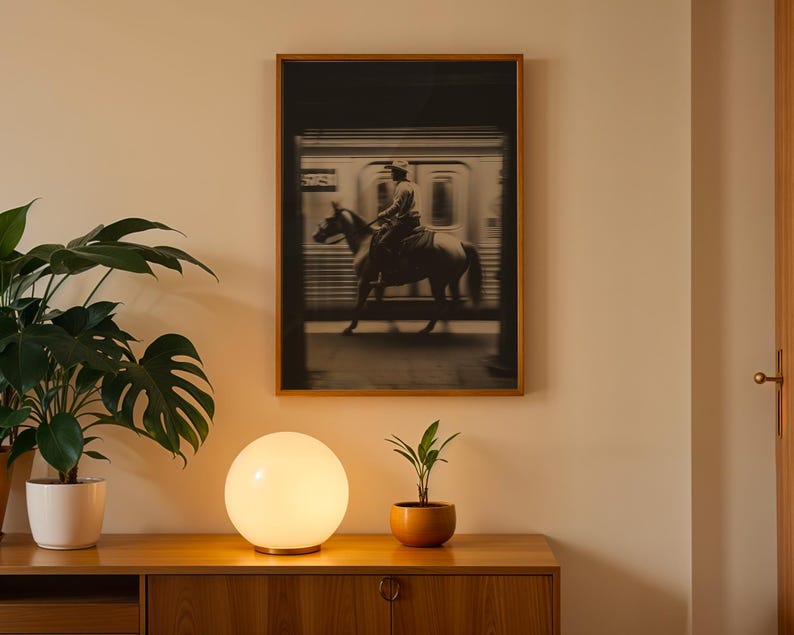 May include: A framed black and white photograph of a person riding a horse near a train. The photograph is mounted on a light-colored wall above a wooden cabinet. A glowing white orb lamp and potted plants are on the cabinet.