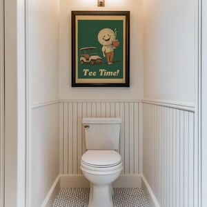 May include: A white toilet with a white and black tiled floor in a bathroom with white wainscoting. A framed poster with a green background and a cartoon golf ball character holding a cup of coffee and riding in a golf cart. The poster says "Tee Time!"