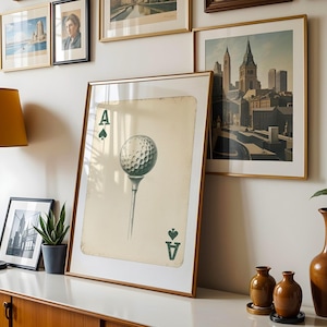 May include: A framed print of an ace of spades playing card with a golf ball on a tee in the center. The card is on a white background with a light brown wood frame.