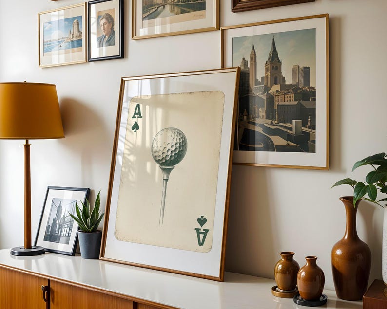May include: A framed print of an ace of spades playing card with a golf ball on a tee in the center. The card is on a white background with a light brown wood frame.